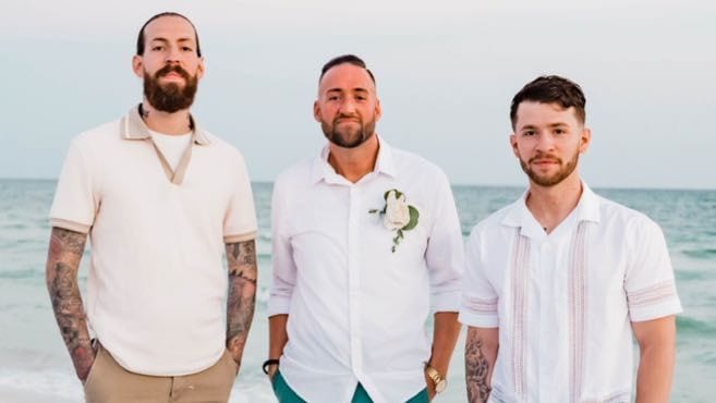 Three men in white and cream shirts standing side by side on a beach, the sea behind them.